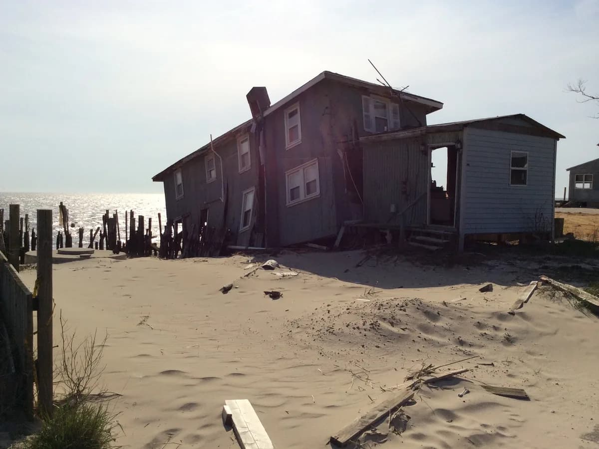 A storm-damaged house half buried in sand at the water line